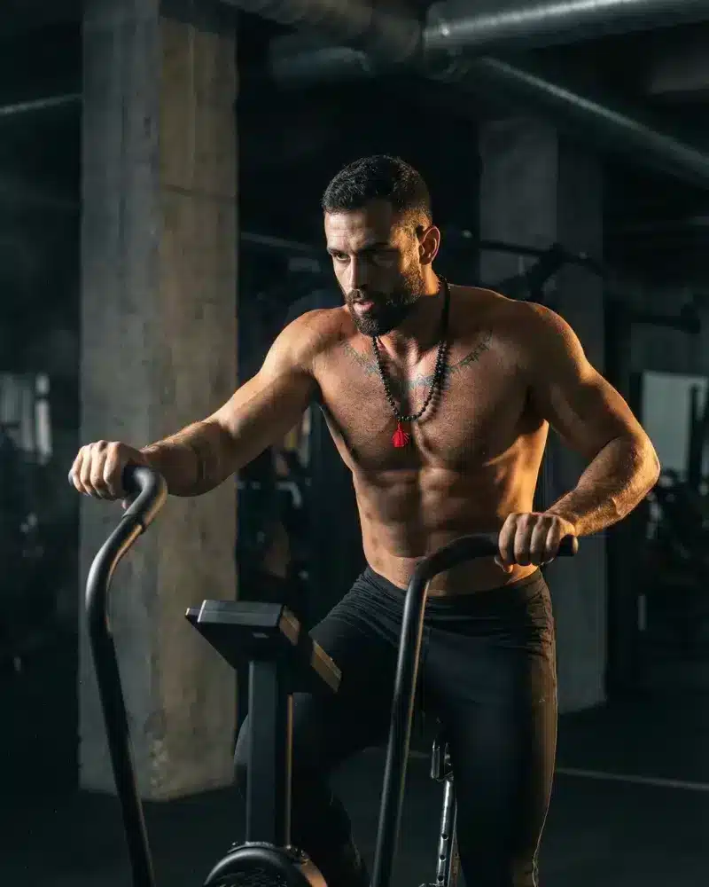 A fit man performs a vigorous, full-body workout on an assault bike inside a gym setting.